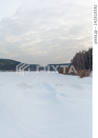 Snow-covered river landscape features trees lining the banks under a cloudy sky, creating a serene winter scene with a frozen surface 142818592