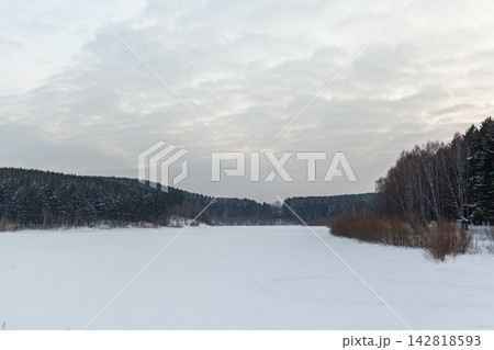 Snow-covered river landscape features a dense forest and cloudy sky, creating a serene winter scene with a frozen water surface 142818593