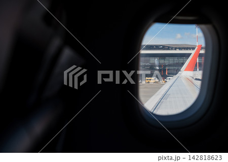 Airplane wing visible from cabin window with airport terminal and ground vehicles seen outside, capturing a moment during daytime travel Airplane wing visible from cabin window with airport terminal and ground vehicles seen outside, capturing a moment during daytime travel 142818623