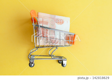 Mini shopping cart contains 5000 ruble banknotes against a vibrant yellow background, illustrating concepts of shopping and finance 142818629