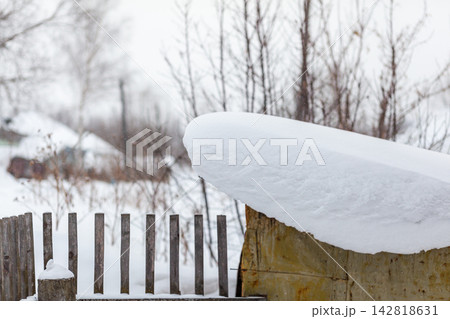 Snow blankets a wooden fence and shed in a rural area, with leafless trees and distant houses creating a serene winter scene 142818631
