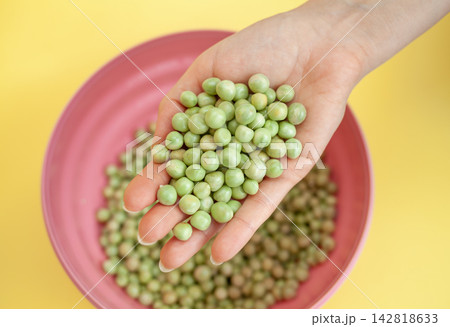 Hand displays fresh green peas above a pink bowl filled with additional peas, set against a vibrant yellow background Hand displays fresh green peas above a pink bowl filled with additional peas, set against a vibrant yellow background 142818633