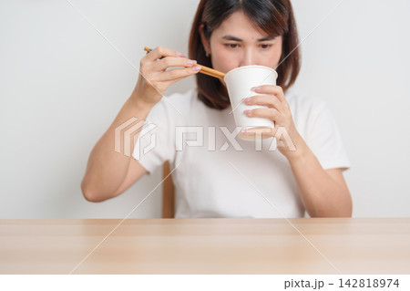 Happy young Asian woman enjoying a delicious cup of hot instant noodles while relaxing at home, Cheerful female eating instant ramen with a smile at apartment. Simple lifestyle and quick meal concept 142818974