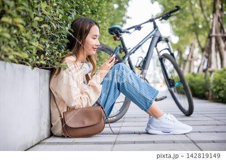 A cute young woman seated with her bicycle enjoys the beauty of a spring day in the park. She listens to music with earphones and types a joyful message on her mobile phone reflecting her happiness. A cute young woman seated with her bicycle enjoys the beauty of a spring day in the park. She listens to music with earphones and types a joyful message on her mobile phone reflecting her happiness. 142819149