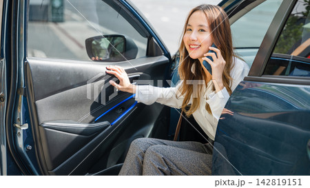 In the urban landscape, an Asian businesswoman stands beside her car, holding a phone and an open door. She exudes confidence and success as she manages business calls while on the move. In the urban landscape, an Asian businesswoman stands beside her car, holding a phone and an open door. She exudes confidence and success as she manages business calls while on the move. 142819151