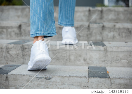 A close-up of a determined young woman's legs in sneakers as she ascends a modern city stairway. Each stepping motion symbolizes her unwavering determination, progress, and the path to success. A close-up of a determined young woman's legs in sneakers as she ascends a modern city stairway. Each stepping motion symbolizes her unwavering determination, progress, and the path to success. 142819153