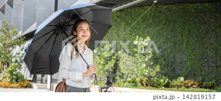 A businesswoman enjoys a sunny countryside ride on her bicycle protected by her umbrella. Her radiant smile and carefree demeanor symbolize the joy of outdoor freedom and the beauty of nature seasons. A businesswoman enjoys a sunny countryside ride on her bicycle protected by her umbrella. Her radiant smile and carefree demeanor symbolize the joy of outdoor freedom and the beauty of nature seasons. 142819157