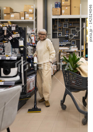 Senior woman smiling, holding a vacuum cleaner while shopping in a retail store Senior woman smiling, holding a vacuum cleaner while shopping in a retail store 142824046