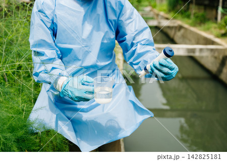 Environmental researcher in blue gown testing water purity in a beaker using a digital meter 142825181