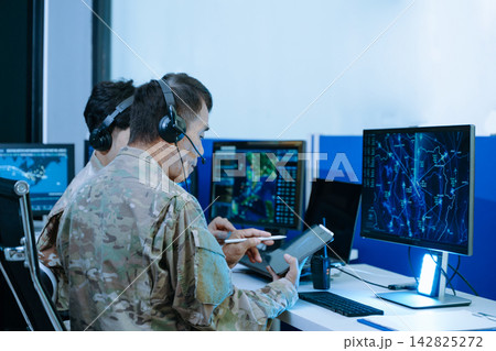 Senior military officer in camouflage using a tablet to analyze tactical radar data alongside his team in a modern cybersecurity command center. 142825272