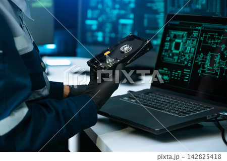 engineer in mask and gloves analyzing an open hard drive for data recovery. Futuristic cybersecurity laboratory with glowing digital screens 142825418