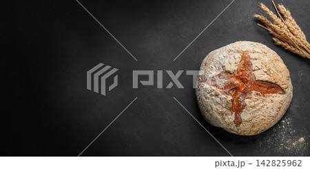 Round artisanal bread loaf with a cross pattern on a dark surface, accompanied by wheat stalks, showcasing rustic baking and natural ingredients. copy space Round artisanal bread loaf with a cross pattern on a dark surface, accompanied by wheat stalks, showcasing rustic baking and natural ingredients. copy space 142825962