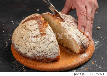 Freshly baked round bread loaf being sliced with a knife on a wooden cutting board, flour dust visible on the surface and around the loaf 142825971