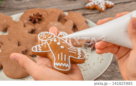 Gingerbread cookie being decorated with white icing by a person holding a piping bag, with additional cookies and star anise on a wooden table 142825985