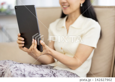 ソファに座りながら笑顔でタブレットを操作する女性の手元のクローズアップ 142827902