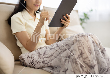 ソファに座りながらペンを使ってタブレットを操作する女性の手元のクローズアップ 142828202