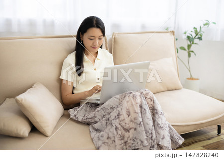 ソファに座りながらノートパソコンを操作する若い笑顔の日本人女性 ソファに座りながらノートパソコンを操作する若い笑顔の日本人女性 142828240