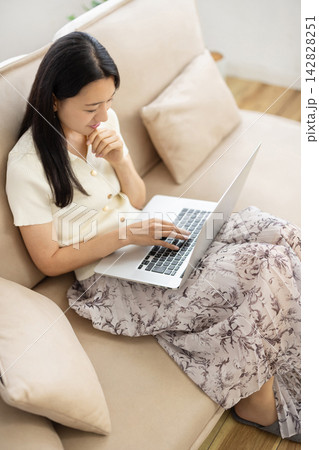 ソファに座りながらノートパソコンを操作する若い笑顔の日本人女性 142828251