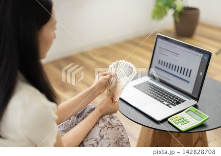 ノートパソコンを見ながら日本円の札束を数える女性の後姿 ノートパソコンを見ながら日本円の札束を数える女性の後姿 142828708