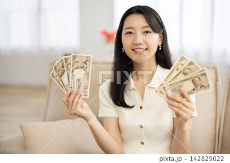 ソファに座り日本円の札束を持ってカメラ目線で微笑む若い日本人女性 ソファに座り日本円の札束を持ってカメラ目線で微笑む若い日本人女性 142829022