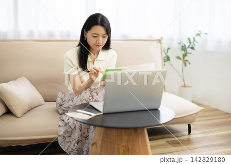 ノートパソコンと通帳と一万円札がのったテーブルの前で電卓を使う若い日本人女性 142829180