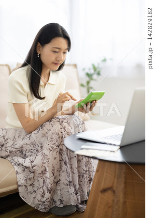 ノートパソコンと通帳と一万円札がのったテーブルの前で電卓を使う若い日本人女性 142829182