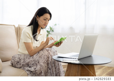 ノートパソコンと通帳と一万円札がのったテーブルの前で電卓を使う若い日本人女性 142829183
