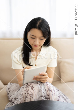 窓辺のソファでノートパソコンを見ながらノートにメモをする若い日本人女性 142829348