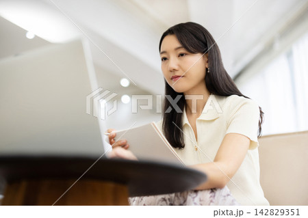 窓辺のソファでノートパソコンを見ながらノートにメモをする若い日本人女性 窓辺のソファでノートパソコンを見ながらノートにメモをする若い日本人女性 142829351