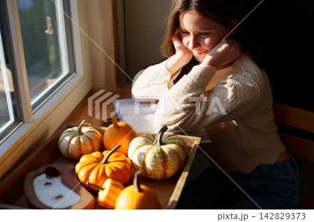 Teen girl relaxing by window with decorative pumpkins at home Teen girl relaxing by window with decorative pumpkins at home 142829373