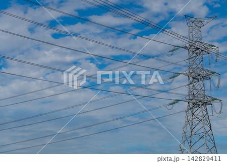 High voltage electricity transmission tower and power lines against blue cloudy sky, electrical grid infrastructure delivering energy for urban communities, industry and modern power distribution. High voltage electricity transmission tower and power lines against blue cloudy sky, electrical grid infrastructure delivering energy for urban communities, industry and modern power distribution. 142829411