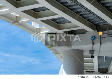 CCTV surveillance camera installed beneath a concrete and steel building structure, monitoring an outdoor area during daytime for security, safety, property protection and crime prevention. 142829415