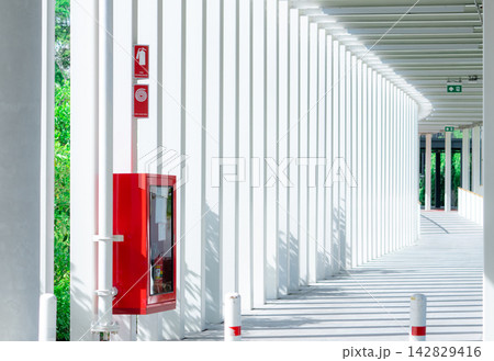 Red fire extinguisher cabinet along an empty modern building corridor with repeating white columns and sunlight shadows, providing emergency fire safety equipment for public building protection. 142829416
