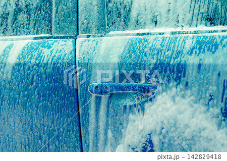 Blue car door covered with thick white soap foam during vehicle washing, automotive cleaning and maintenance service for removing dirt before exterior detailing, polishing and protective waxing. Blue car door covered with thick white soap foam during vehicle washing, automotive cleaning and maintenance service for removing dirt before exterior detailing, polishing and protective waxing. 142829418