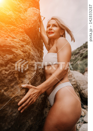 Fit blonde woman in white bikini poses against large rock formation outdoors during sunny day 142833287