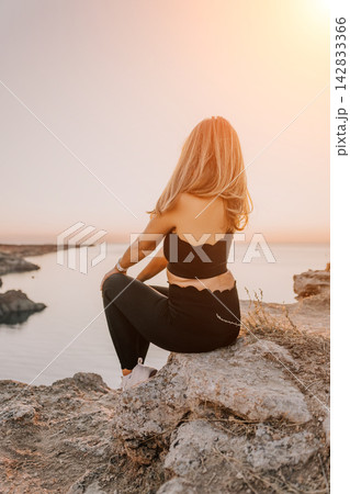 Woman sunset cliff sits on a rocky ledge overlooking the ocean during a peaceful golden hour sunset for meditation and relaxation 142833366