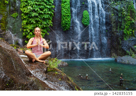 Young woman meditating in yoga pose under Banyumala Twin Waterfalls. Water flowing under cascading high waterfall.  Adventure lifestyle and retreat at holidays travel in Bali tropical rainforest. 142835155