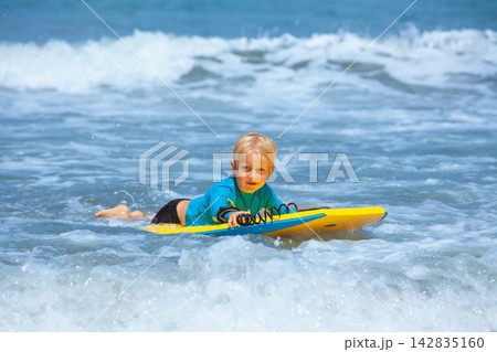 Happy young surfer boy with bodyboard have fun on beach, riding on small ocean waves. Summer adventures, active families, parents and kids lifestyle, sports activities on school holiday with kids Happy young surfer boy with bodyboard have fun on beach, riding on small ocean waves. Summer adventures, active families, parents and kids lifestyle, sports activities on school holiday with kids 142835160