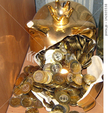 Broken golden piggy bank with a crown overflowing with metallic coins on a wooden shelf, symbolizing savings, wealth, and financial concepts. 142835538