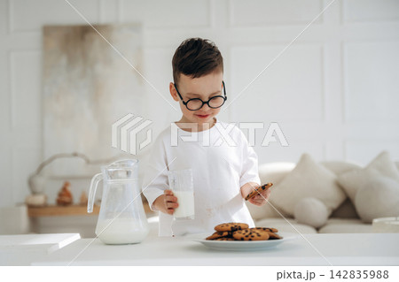 Young boy with glasses holding cookie and glass of milk in bright kitchen, plate of chocolate chip cookies on table with soft furnishings in the background Young boy with glasses holding cookie and glass of milk in bright kitchen, plate of chocolate chip cookies on table with soft furnishings in the background 142835988