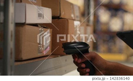 African american woman using scanner to register barcodes tags in inventory, solving logistics operations in industrial warehouse. Worker checking shipping labels near storage racks. Camera B. African american woman using scanner to register barcodes tags in inventory, solving logistics operations in industrial warehouse. Worker checking shipping labels near storage racks. Camera B. 142836140