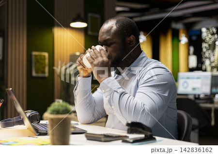 African american male analyst drinking coffee cup to keep working at night, staying awake to finish administrative tasks in the office. Sipping on a caffeine drink after hours. African american male analyst drinking coffee cup to keep working at night, staying awake to finish administrative tasks in the office. Sipping on a caffeine drink after hours. 142836162