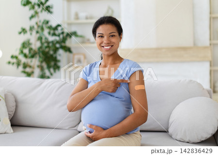 Pregnant Black woman at home, pointing to bandage on arm after vaccination Pregnant Black woman at home, pointing to bandage on arm after vaccination 142836429