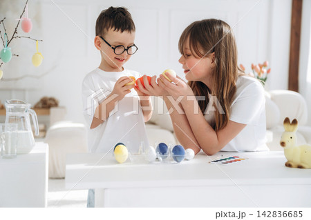 Two children, a boy and a girl, wearing white shirts, play with colorful Easter eggs at a bright table in a well-lit room decorated for spring 142836685