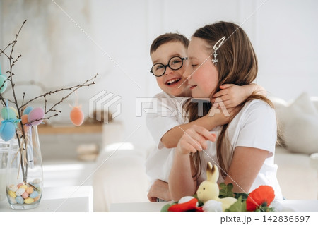 Young boy in glasses hugs older sister while sitting at a table decorated with Easter eggs and flowers in a bright, modern indoor setting 142836697