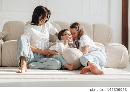 Woman and two children sitting on a light-colored rug in a bright living room, engaging in playful interaction while seated on a comfortable sofa 142836958