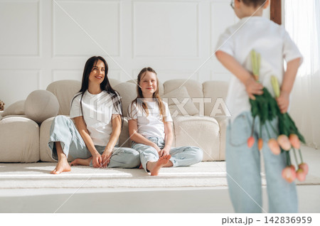 Young boy holding bouquet of tulips behind his back surprises mother and sister sitting on a beige sofa in a bright, modern living room with white walls 142836959