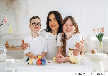Woman with two children wearing white shirts and bunny ears, celebrating Easter with colorful eggs and decorations in a bright, modern living room Woman with two children wearing white shirts and bunny ears, celebrating Easter with colorful eggs and decorations in a bright, modern living room 142836973