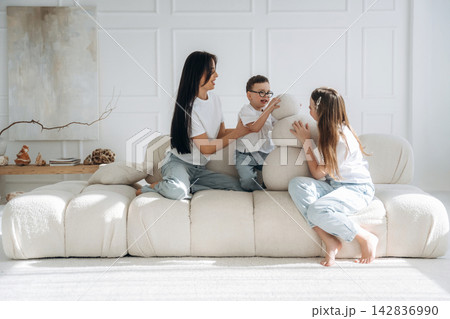 Family of three enjoying playful moments on a modern white sofa in a bright living room with minimalistic decor and natural light streaming through large windows Family of three enjoying playful moments on a modern white sofa in a bright living room with minimalistic decor and natural light streaming through large windows 142836990