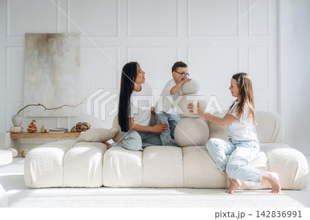 Two women and a boy play with a soft sculpture on a modern white sofa in a bright, minimalist living room with natural light and decorative elements Two women and a boy play with a soft sculpture on a modern white sofa in a bright, minimalist living room with natural light and decorative elements 142836991